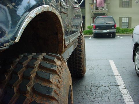 1989 Ford Bronco - A close-up view of a heavily mud-splattered off-road tire and fender, with other vehicles visible in the background.
