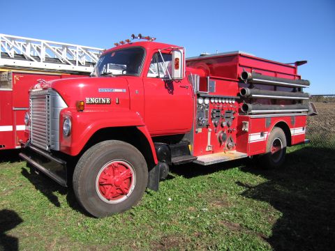 1991 Ford LTD Crown Victoria - A red fire truck with a ladder is parked on a grassy field under a clear blue sky.