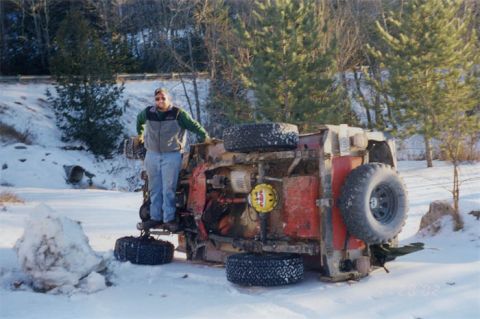 1985 Ford Bronco - An off-road vehicle has flipped onto its side in a snowy landscape, with a person standing nearby.