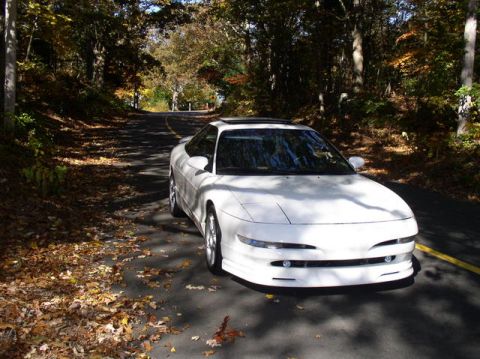 1996 Ford Probe - A white 1996 Ford Probe GT drives down a road lined with trees in autumn.