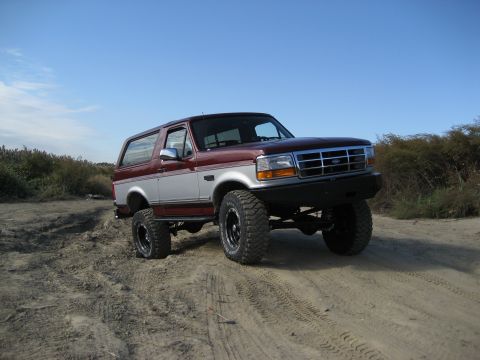 1996 Ford Bronco - A lifted Ford Bronco with two-tone paint sits on a sandy track under a clear blue sky.