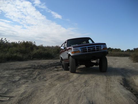 1996 Ford Bronco - A two-tone Ford Bronco drives on a dusty trail under a partly cloudy sky.
