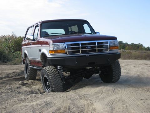 1996 Ford Bronco - Maroon and silver Ford Bronco with aftermarket bumper and large tires stuck in sand.