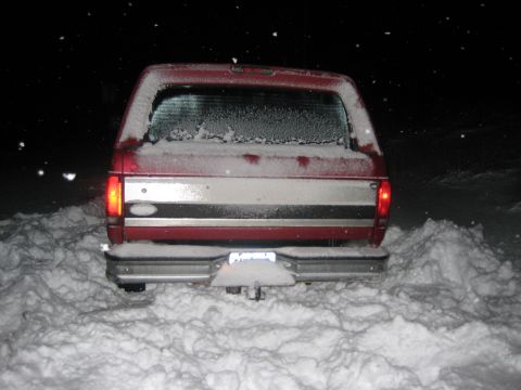 1994 Ford Bronco - A red SUV is partially buried in snow, with its rear lights illuminated.