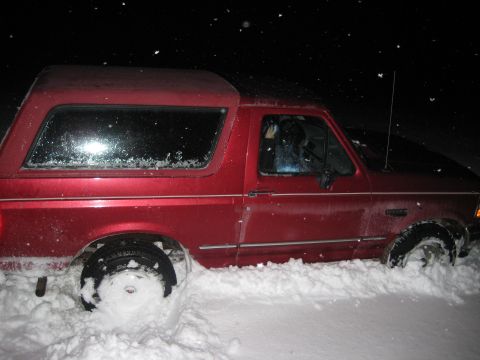 1994 Ford Bronco - A red Ford Bronco is stuck in the snow at night, with snow covering its wheels and lower body.