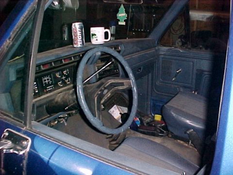 1985 Ford Bronco - The interior of a blue vehicle shows a dashboard with gauges, a steering wheel, and a driver's seat, along with a beverage can and mug.