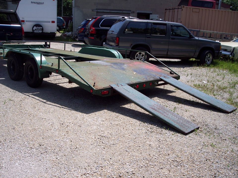 Green car trailer with ramps extended, parked on gravel. SUVs and RV visible in background