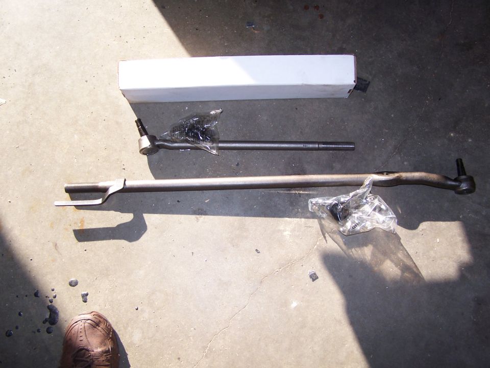 Steering linkage parts with a white box on a concrete surface. A shoe is visible in the lower left