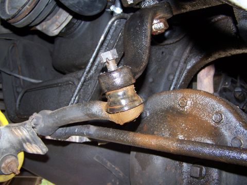 1986 Ford Bronco - Underbody close-up of the drag link tie rod end being separated at the pitman arm, with the nut backed off up the exposed stud and a torn t…