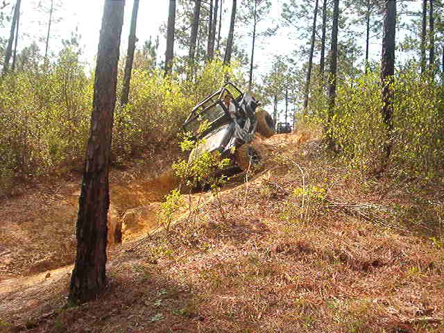 Dark topless Bronco with roll cage climbing a steep dirt trail through pine woods