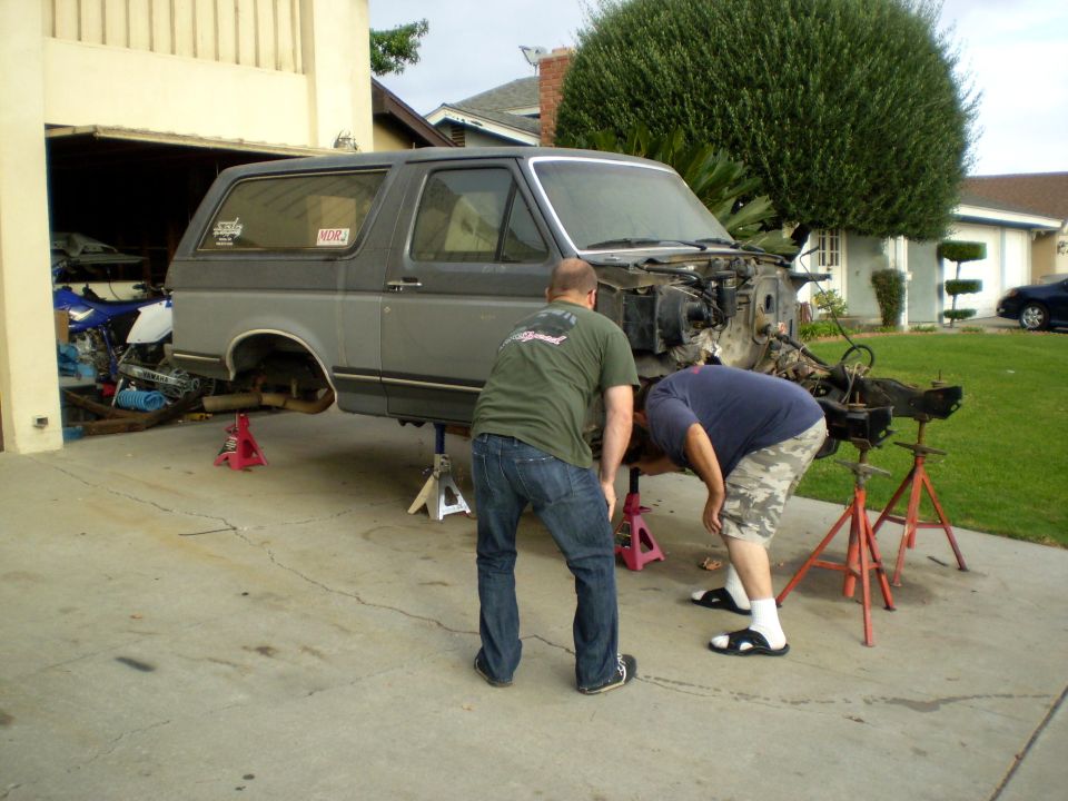Two men working on the chassis of a gray Ford Bronco on jack stands. A dirt bike is in the garage behind.