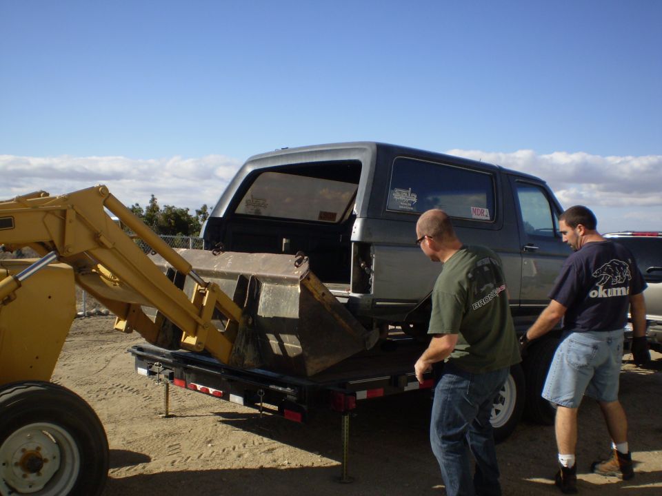 Dark SUV being loaded onto a trailer by a yellow front-end loader, with two men nearby.