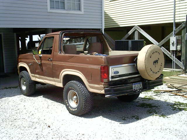 Brown Ford Bronco with top removed, rear 3/4 view, tan Ford Bronco spare tire cover, parked on gravel