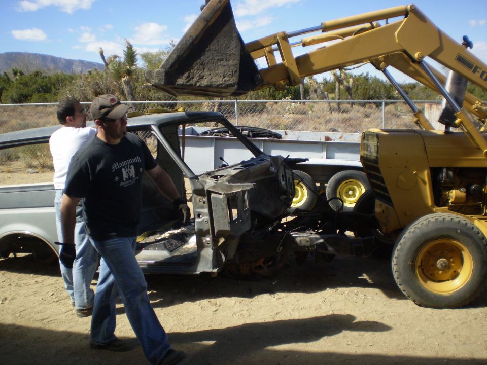 Yellow front-end loader lifting vehicle chassis with two men nearby.