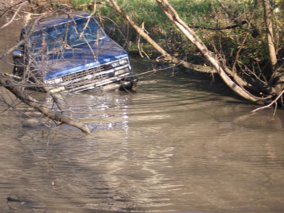 Blue pickup truck stuck in water with branches covering it.