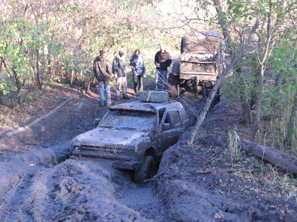 Muddy SUV stuck in a rut on a dirt trail, surrounded by trees, with people watching.