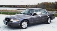 2003 Ford Crown Victoria - A dark gray 2003 Ford Crown Victoria sedan is parked on a gravel surface with a body of water and trees in the background.