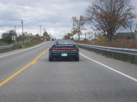 1996 Ford Probe - The rear view of a dark green 1996 Ford Probe is shown driving on a two-lane road. The car has a spoiler and a visible license plate.