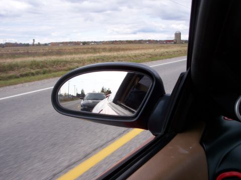 1996 Ford Probe - A side mirror view shows a black car driving on a road behind a red car. The background features a rural landscape with fields and a silo.