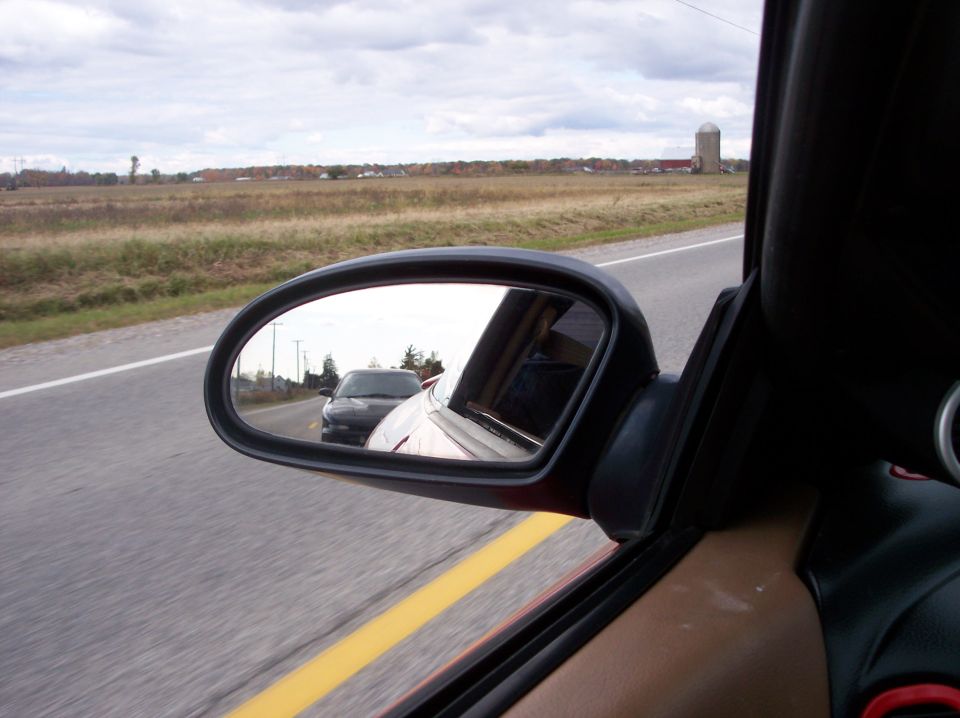 Reflection in side mirror of a black car following a red car on a road, rural background.