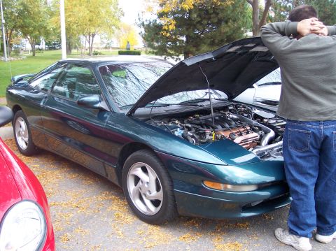 1996 Ford Probe - A dark green 1996 Ford Probe with its hood open is parked on a street covered in fallen leaves. A person stands behind the car, looking at…