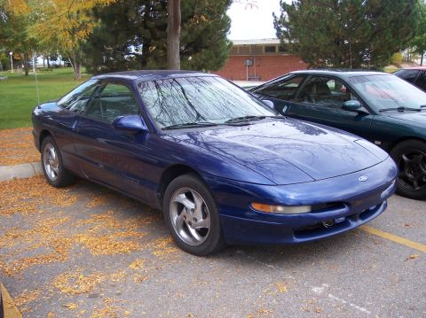 1996 Ford Probe - A blue 1996 Ford Probe is parked on asphalt covered with fallen leaves. A dark green car is parked behind it.