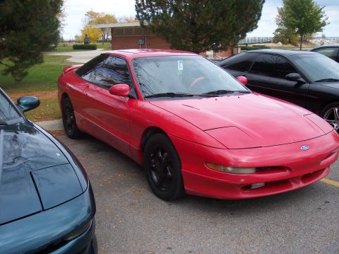 1996 Ford Probe - A red 1996 Ford Probe is parked on asphalt next to a dark green vehicle. The red car is a two-door coupe with black wheels and a spoiler.
