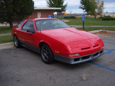 1996 Ford Probe - A red Dodge Daytona coupe is parked in a parking lot with a building and playground in the background. The car features a gray lower body k…