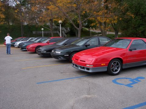 1996 Ford Probe - A line of cars parked in a lot, with a red sports coupe in the foreground. A person in a white t-shirt and jeans stands to the left, facing…