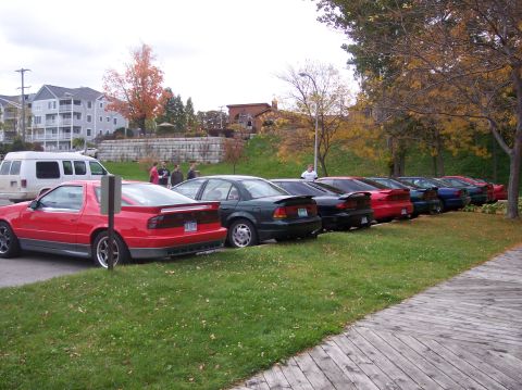 1996 Ford Probe - A line of sports cars is parked on a grassy area next to a wooden boardwalk. The closest vehicle is a red sports coupe with a spoiler, foll…