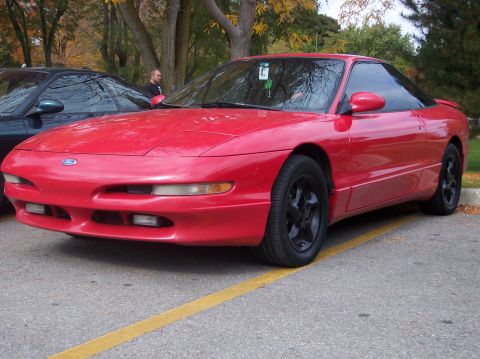 1996 Ford Probe - A red 1996 Ford Probe is parked in a lot next to a dark-colored car. The front of the red sports coupe is visible, showing its headlights,…