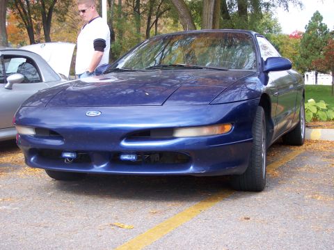 1996 Ford Probe - A front 3/4 view of a dark blue 1996 Ford Probe parked on asphalt with fallen leaves scattered around. The car has aftermarket blue lights…