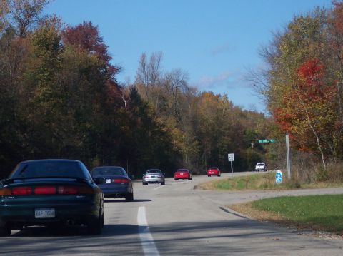 1996 Ford Probe - A dark green 1996 Ford Probe is shown from the rear, driving on a road with autumn foliage on either side. Other cars are visible ahead on…