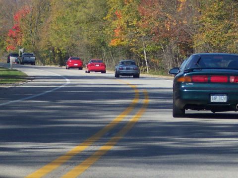 1996 Ford Probe - The rear of a dark green 1996 Ford Probe with a spoiler is visible on a two-lane road during autumn. The car has a license plate that reads…