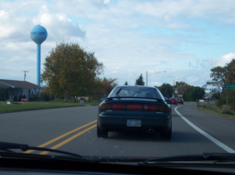 1996 Ford Probe - The rear of a dark green 1996 Ford Probe is visible driving on a road with double yellow lines. A blue water tower labeled "PORT HOPE" is i…