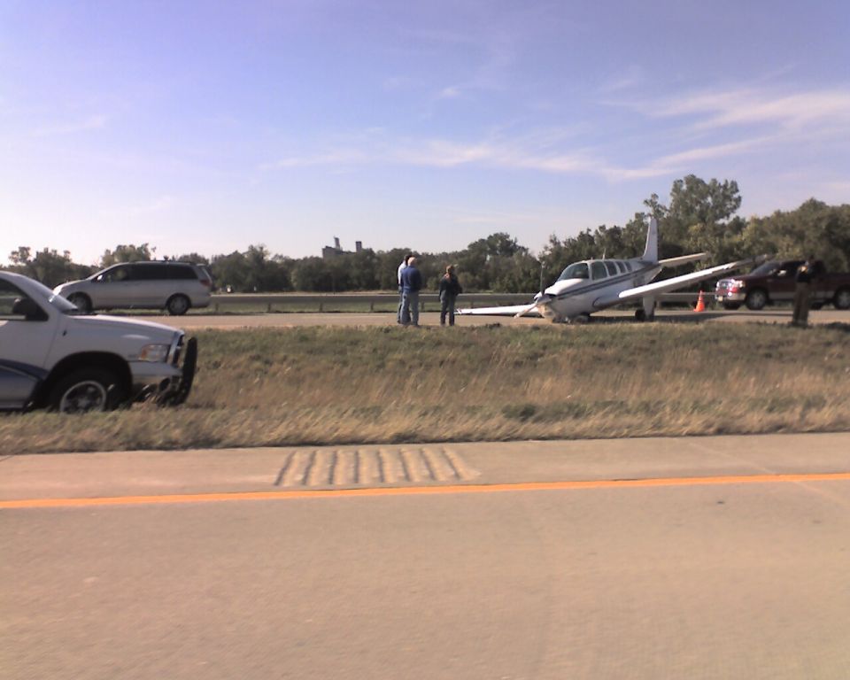 Small airplane on the side of a highway with people and vehicles.