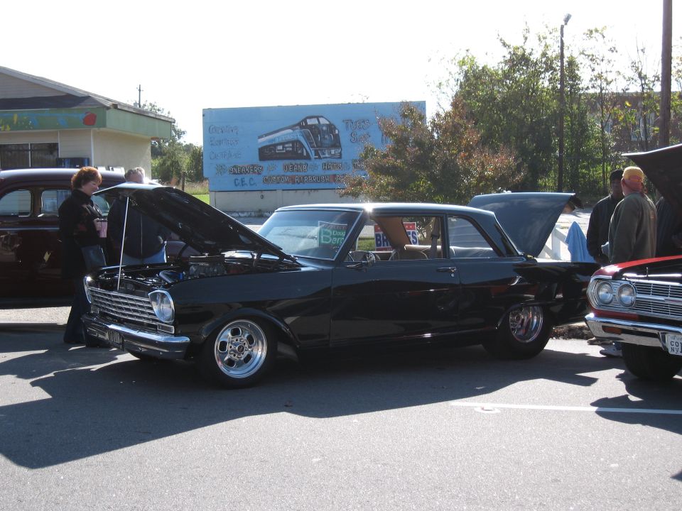 Black classic coupe with hood open, chrome trim, custom wheels, parked on asphalt. Red classic car visible.