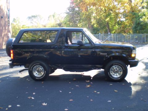 1996 Ford Bronco - The side profile of a black 1996 Ford Bronco is shown parked on an asphalt surface with fallen leaves. The vehicle features a spare tire mo…