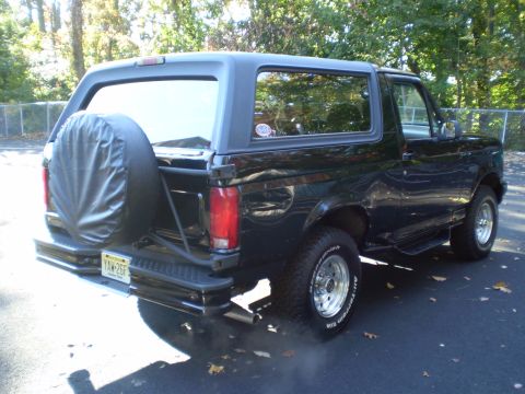 1996 Ford Bronco - The rear driver's side quarter view of a black 1996 Ford Bronco is shown parked on an asphalt driveway. A spare tire covered with a black v…