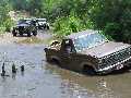 1985 Ford Bronco - Several off-road vehicles navigate a flooded dirt path surrounded by lush greenery.