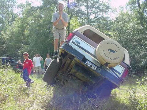 1985 Ford Bronco - A man stands atop a 1985 Ford Bronco that has tipped onto its side, with other people and vehicles in the background.