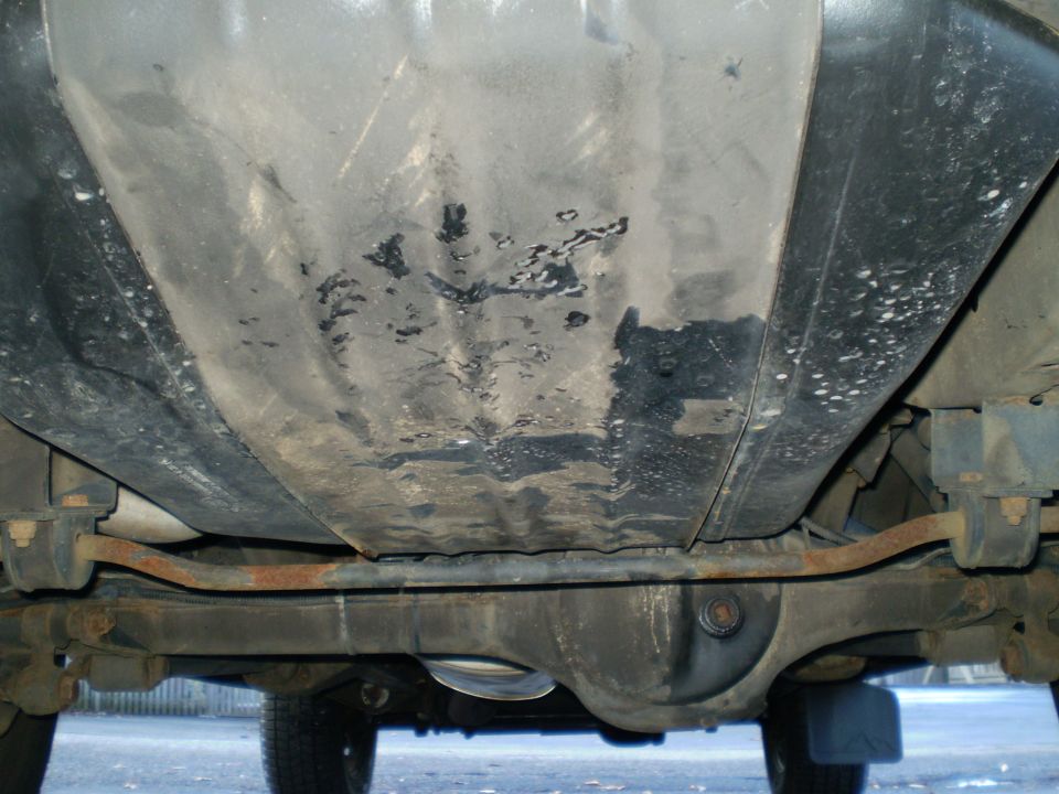 Underbody view of a grimy fuel tank and surface-rusted anti-roll bar beneath a vehicle