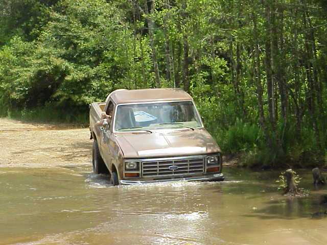Tan Ford Bronco with doors removed driving through muddy water at a wooded pond
