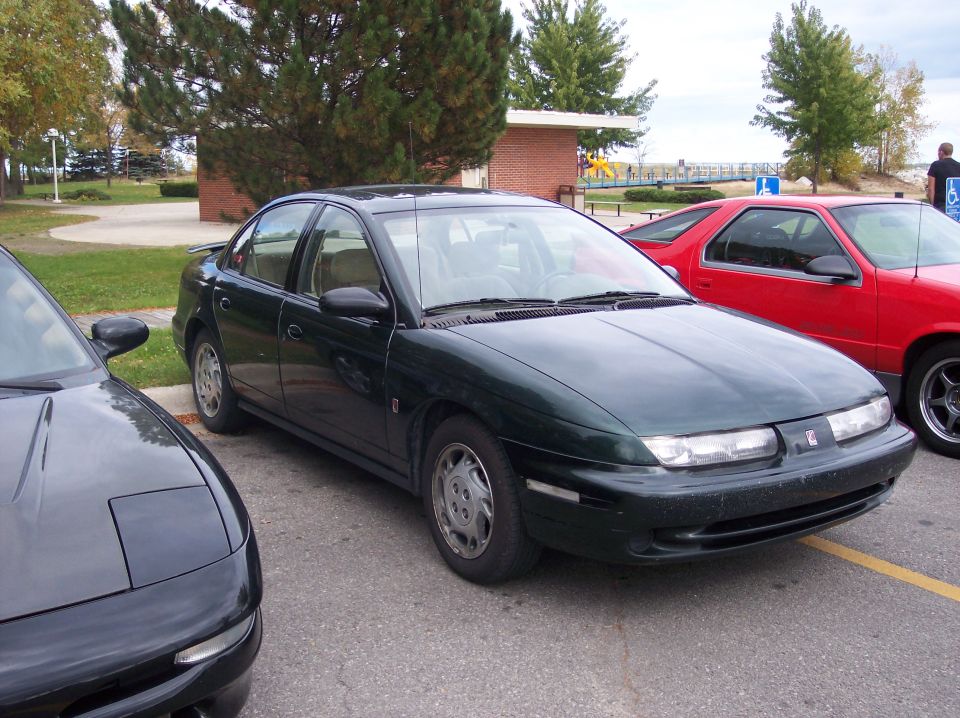 Dark green Saturn sedan parked in a lot next to a black sports car and a red car.