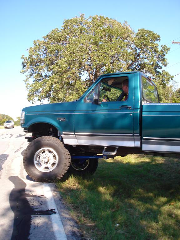 Teal Ford F-150 pickup truck with large tires and lifted suspension parked on the side of the road.