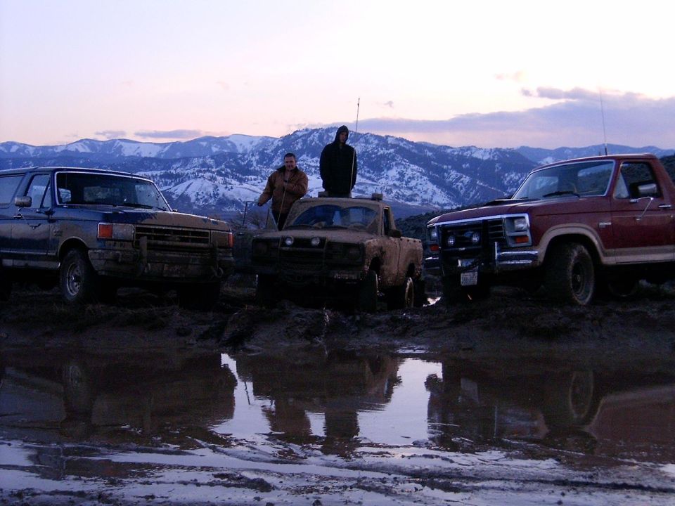 Three muddy Ford Bronco SUVs stuck in a muddy field with mountains in the background.