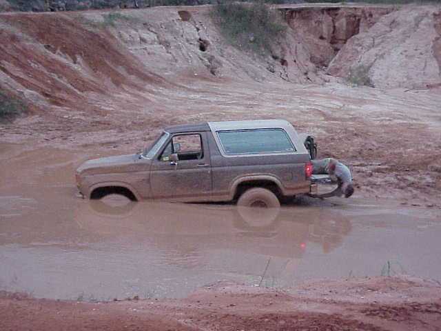Muddy Ford Bronco stuck axle-deep in a clay pit while a man hooks a strap to the rear