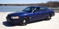 2003 Ford Crown Victoria - A dark blue Ford Crown Victoria sedan is parked on a concrete surface next to a body of water. The car is shown from a three-quarter front…