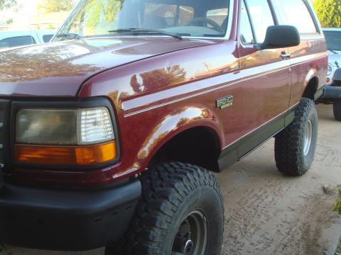 1992 Ford Bronco - A close-up, low-angle shot of the front passenger side of a dark red Ford Bronco. The vehicle features large, off-road tires and a black bu…