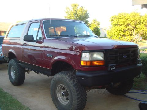 1992 Ford Bronco - A maroon Ford Bronco is parked on a concrete driveway in front of a house with trees in the background. The vehicle has large off-road tire…